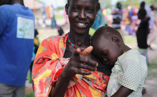 En kvinne peker på kamera og holder sin sovende baby mens familier står i kø for å bli registrert for hjelp og matutdeling i Sør-Sudan.