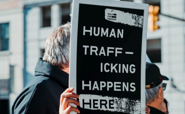 Antall ofre for menneskehandel som ble oppdaget rundt om i verden falt i 2020 med 11 prosent fra året før, ifølge en ny UNODC -rapport. Her står en mann med et skilt hvor det står: «Human trafficking happens here», eller «Menneskehandel skjer her» for å sette fokus på menneskehandel som et globalt problem i dag. Foto: Hermes Rivera/Unsplash.