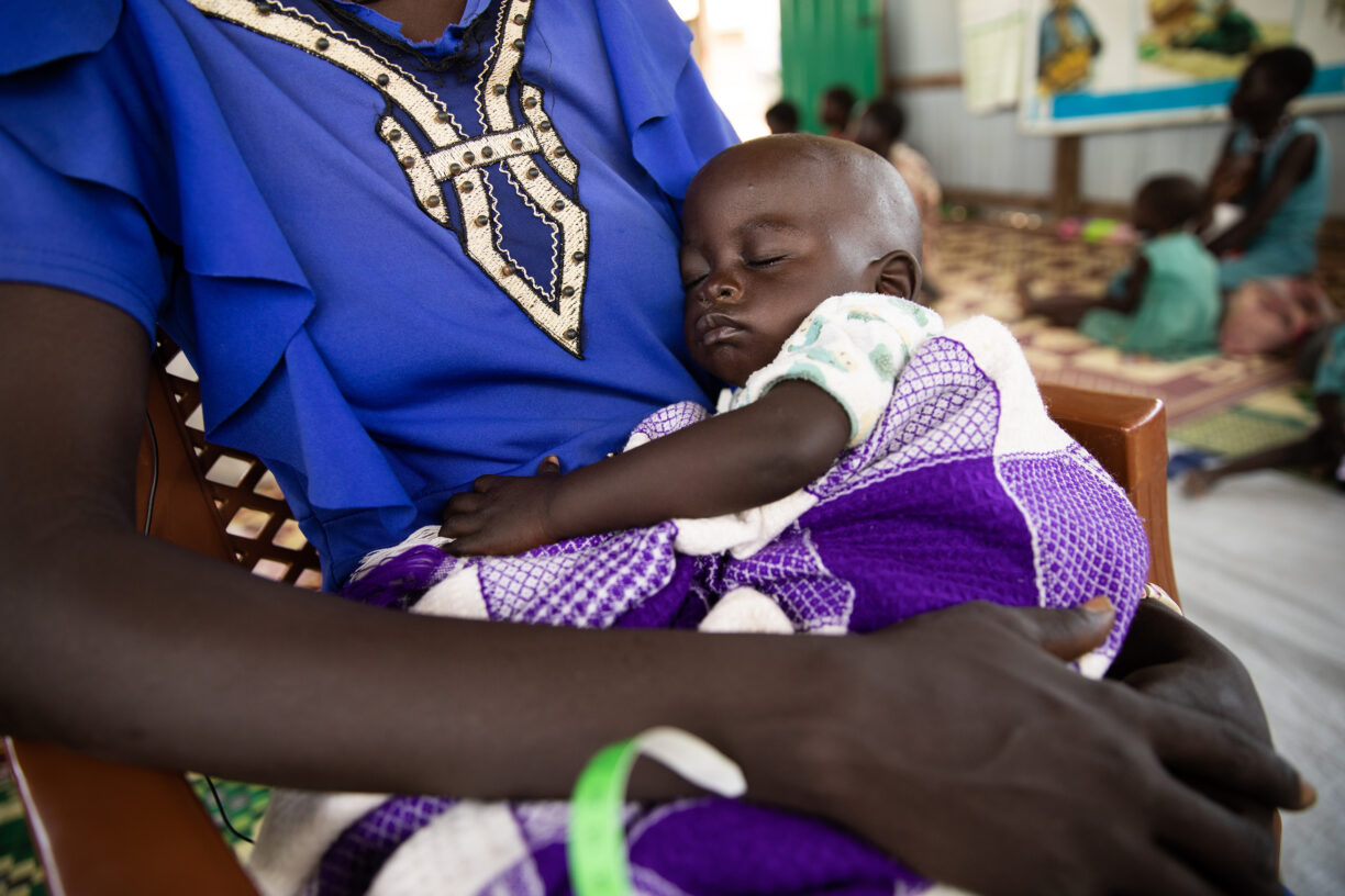 På dette bildet venter Adut (28) og baby Achan (1½) på å bli sett av det medisinske personalet ved ernæringssenteret i Alek, Sør-Sudan. Adut hadde en komplisert graviditet og fødsel, og Achan har mottatt kosttilskudd mot underernæring fra WFP siden han var seks måneder gammel. Sammen må Adut og baby Achan gå én time hver vei, til og fra senteret, og de er på senteret jevnlig for kontroll og for å få kosttilskudd. Foto: WFP/Samantha Reinders.